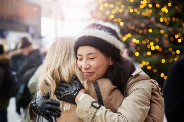 Asian woman embraces friend in railroad station, christmas tree in background.